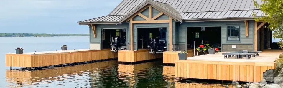 Wooden boathouse on a lake, with a clean, modern metal roof. Boats are visible inside through the three open doorways. The boathouse is surrounded by water and a rocky shoreline.