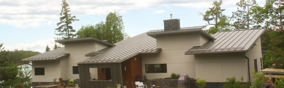 Modern single-story house with a series of overlapping metal roofs, surrounded by trees. There is a large wooden deck with patio furniture and potted plants. There is a lake in the background.