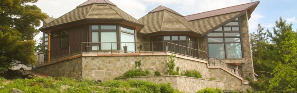 A modern house, with large glass windows, stone walls, and a unique angular roof design. The roof features sections of modern-style and shingle-style metal roofing. The house has a balcony with metal railing and is surrounded by lush greenery.