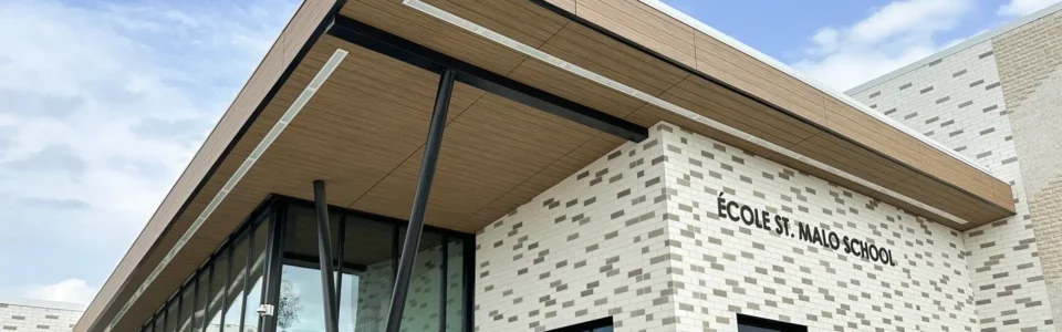 Close-up of Ecole St. Malo School in Saint Malo, Manitoba. The school is a modern building featuring a patterned brick wall with light and dark bricks, and large glass windows in the entryway. The overhanging roof has wood-patterned metal fascia and soffits that contrast with the brick.