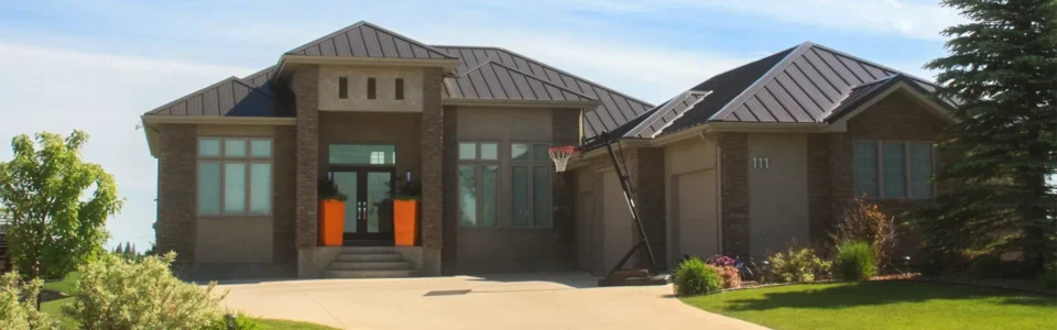 A large modern house, featuring a standing seam metal roof. The roof is dark brown, with clean, parallel lines. The house has a stone exterior and an attached garage, with a wide concrete driveway. The front yard is well-manicured.