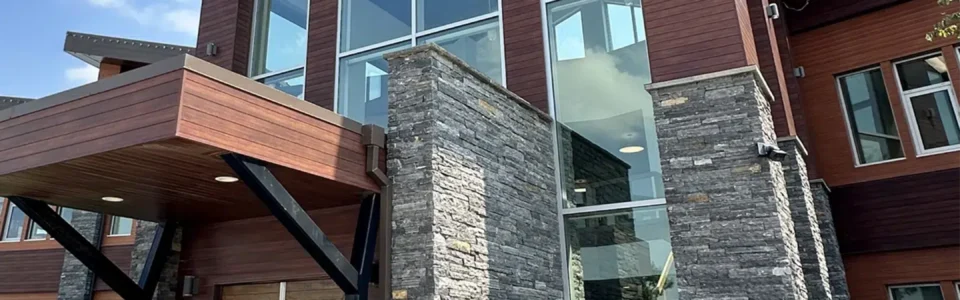 The entryway of a modern two-story building in Winnipeg, Manitoba. The entryway features an overhang with metal siding, soffits, and fascia, along with angular metal roofs, large windows, and brown metal siding that resembles wood. Stone accents frame the front doors.
