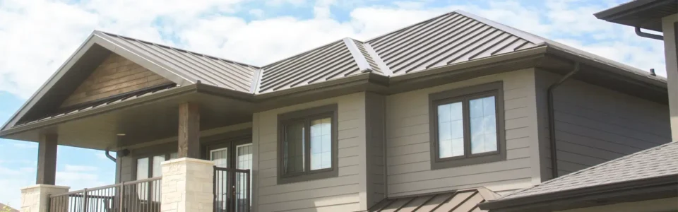 A modern two-story house featuring a brown standing-seam metal roof from Temple Metal Roofs, beige siding, and a covered balcony with wooden and stone pillars.